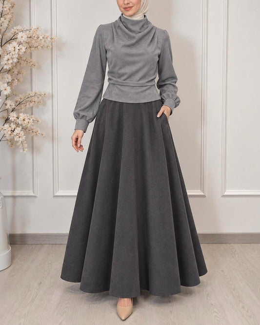 A woman wearing a gray mock-neck long-sleeve top and a matching dark gray flared maxi skirt, paired with a light beige hijab and beige pointed-toe flats, standing in a decorated indoor setting.